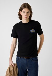 Czarny t-shirt z krótkimi rękawami, z niebiesko-złotym logo "Guess" na piersi. Model nosi niebieskie dżinsy i trzyma beżową kurtkę.