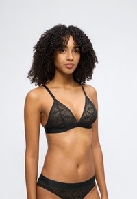 Femme aux cheveux bouclés portant un soutien-gorge en dentelle florale noire et une lingerie assortie, debout devant un fond gris clair uni.