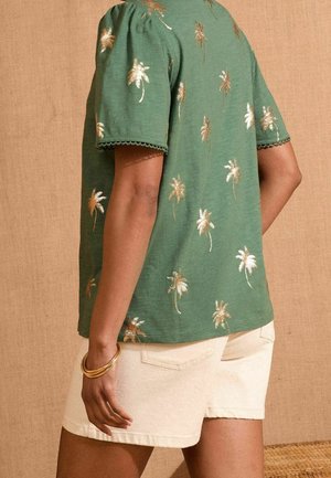 Grøn bluse med korte ærmer med guld palmebladbroderi, bølgede ærmekanter, parret med lyse shorts. Tekstureret baggrund.