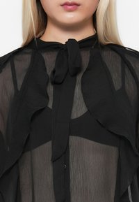 Svart gjennomsiktig bluse med volanger, knyting i halsen og knappelukking. Har en myk, lett tekstur med lagdelte detaljer.