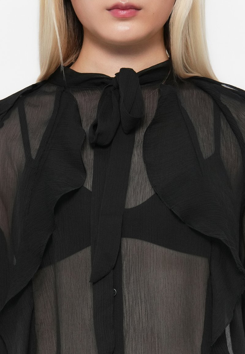Blouse noire transparente avec des détails volantés, un design à nouer au cou et une fermeture à boutons. Présente une texture douce et légère avec des accents superposés.