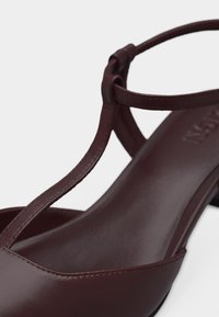 Sandale à talon en cuir brun foncé avec lanière en T et bride de cheville réglable sur fond blanc.