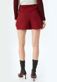 Burgunderfarbene Shorts mit maßgeschneidertem Schnitt, glattem Stoff und zwei Gesäßtaschen. Kombiniert mit grauen Kniestrümpfen und burgunderfarbenen High-Heels.
