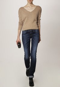 Suéter beige de cuello en V, con silueta ajustada, combinado con jeans skinny azul oscuro. Botines negros y un pequeño bolso de mano redondo. Textura suave.