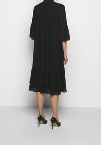 Robe noire avec des manches évasées et transparentes. Présente un ourlet à volants superposés. Le matériau semble léger ; design minimaliste. Portée avec des escarpins noirs.