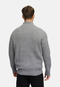 Suéter de punto gris con textura acanalada, cuello alto y mangas largas; presenta un ajuste relajado y un dobladillo ligeramente ajustado.