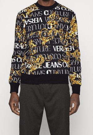 Schwarzes Crewneck-Sweatshirt mit weißem Versace Jeans Couture Schriftzug und goldenem barockem ornamentalen Scrollmuster, getragen mit schwarzer Hose.