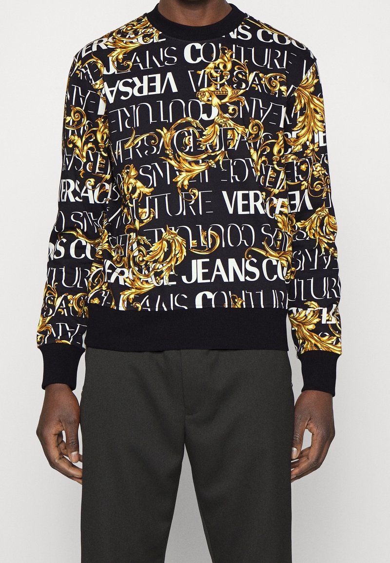 Sweat-shirt noir col rond avec texte blanc Versace Jeans Couture et motif baroque doré orné en volute, porté avec un pantalon noir.