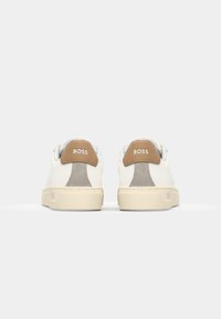 Baskets en cuir blanc avec un accent de talon en daim beige, une semelle en caoutchouc texturée et un logo embossé sur le talon.