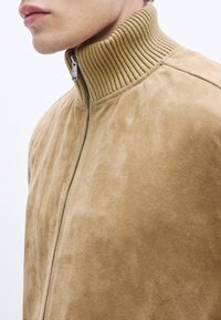 Tan-nahast seemisnahast jakk soonilise rohelise kaelusega, millel on kogu pikkuses tõmblukk ja sile pind, esitades minimalistlikke disainielemente.