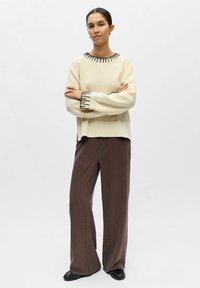 Femme debout, bras croisés, portant un pull crème avec des bordures noires, un pantalon large marron et des chaussures plates noires sur un fond blanc.