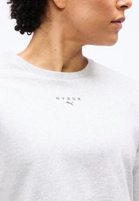 Κοντινό πλάνο του λαιμού και του ώμου ενός ατόμου που φοράει ανοιχτό γκρι φούτερ με την επιγραφή «HYROX» και το λογότυπο της Puma τυπωμένα κάτω από το γιακά.