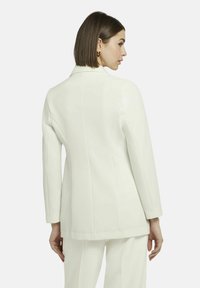 Weißes tailliertes Blazerjackett mit langen Ärmeln und strukturierten Schultern. Die glatte Textur, der einzelne Rückenventil und die klaren Linien unterstreichen das moderne Design.