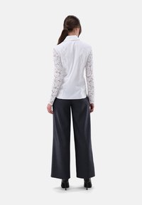 Weißes Bluse mit langen Spitzenärmeln, enganliegendem Rücken, kombiniert mit weit geschnittenen dunklen Hosen und schwarzen High-Heels. Neutraler Hintergrund.