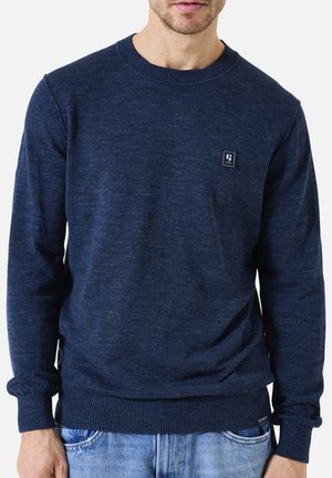 Marineblauwe crewneck trui in een textuurgebreide stof, met een klein logo op de borst en geribbelde mouwen en zoom. Gecombineerd met lichtblauwe jeans.