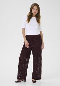Pantaloni larghi in pizzo bordeaux con un motivo floreale testurizzato, abbinati a una maglietta corta bianca aderente. Scarpe scure e piatte completano il look.
