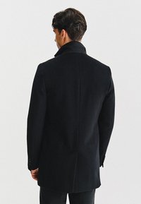 Czarny wełniany płaszcz z wysokim kołnierzem, dopasowany krój, gładka tekstura i guziki na rękawach, pokazany z tyłu na jednolitym tle.