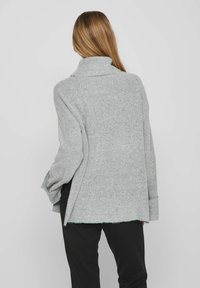 Grauer, strukturierter, oversize Pullover mit hohem Kragen und weiten Ärmeln, ausgestattet mit einem gespaltenen Saum und weichem Stoff. Getragen mit schwarzen Hosen.