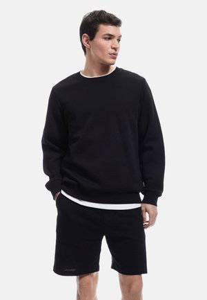 Sweat-shirt noir à col rond avec des poignets côtelés, assorti à un short noir. La tenue comprend une chemise blanche superposée en dessous.