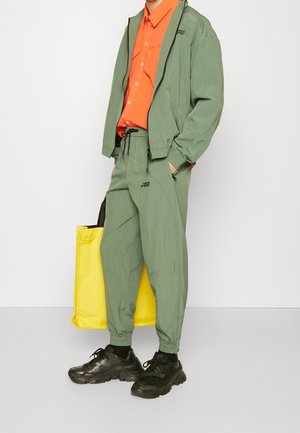 Grön lättviktsjacka och matchande byxor kombinerade med en orange skjorta med knappar och svarta sneakers. En gul tygväska syns.