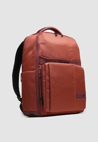 Mochila de tela roja con forma estructurada, múltiples compartimentos con cremallera y un detalle contrastante en marrón en la base. Tirantes acolchados visibles.