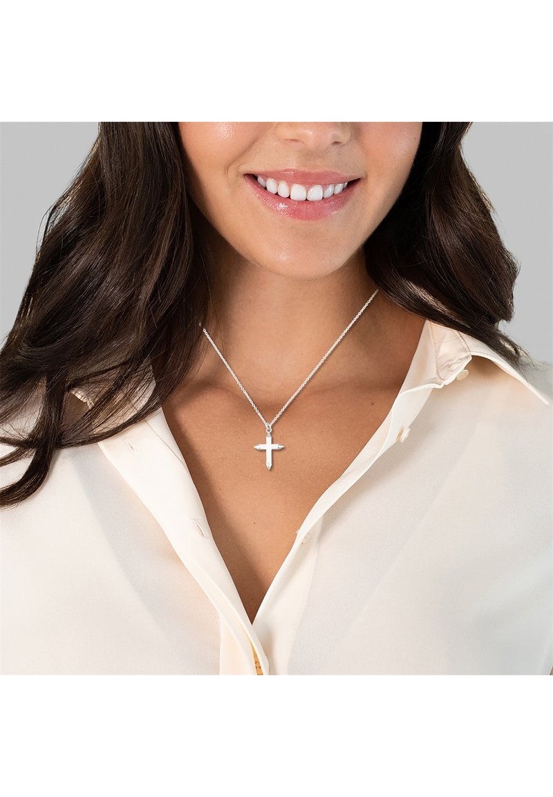 Silverkorssmycke på ett tunt kedja, bärs över en ljusblomblus. Korset har en polerad yta med distinkta armar.