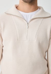 Világos bézs pulóver fél cipzárral, függőleges bordázott textúrával és V-alakú dizájnelemmel a mellkasán. Puha anyagú finish.