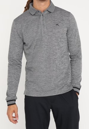 Homme portant un polo gris à manches longues avec col et un pantalon noir, debout avec une main dans sa poche.