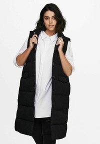 Juodas be rankovių pūkinis liemenė su siuvinėtų raštų dizainu, spaudžių mygtukų užsegimu ir priekinių kišenių, uždėta ant balto marškinėlio.