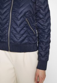 Mörkblå quiltad jacka med chevronmönster, silverdragkedja och ribbade cuffs. Har en sidoficka med dragkedja och slät, glänsande yta.