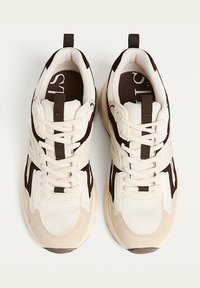 Beige sneakers with brown accents, featuring a mesh upper, round toe, textured surface, and lacing system. Includes a rear pull tab.