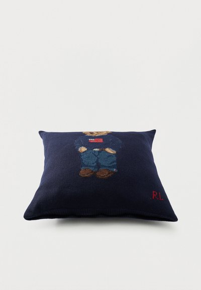 Τετράγωνο μαξιλάρι σε ναυτικό μπλε με πλεκτό αρκουδάκι που φοράει φούτερ με την αμερικανική σημαία, τζιν και καφέ παπούτσια, με κόκκινα αρχικά "RL".