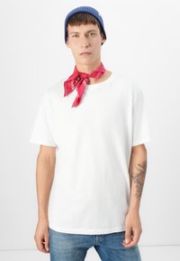 Camiseta blanca de algodón con mangas cortas, combinada con un pañuelo rojo alrededor del cuello y un gorro de canalé azul. Tatuaje distintivo en el brazo.