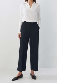 Kvinna klädd i en vit långärmad V-ringad blus, svarta vida capribyxor och svarta spetsiga flatskor, stående med ena handen i fickan.