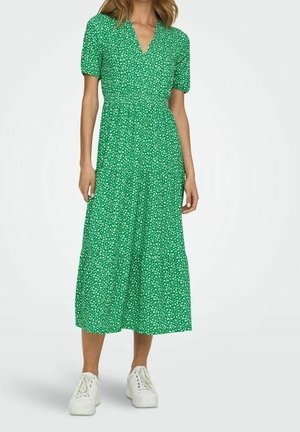 Kvinde iført en grøn midi-kjole med hvidt blomsterprint, korte pufærmer, V-hals og hvide sneakers, stående foran en hvid baggrund.