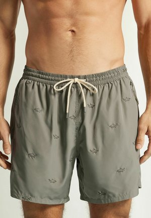 Mand iført olivengrønne badeshorts med hvid snor og små sorte hajtryk, vist fra taljen til midt på låret.