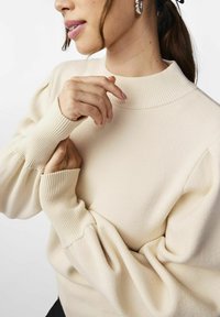 Cremefarbenes Sweater mit hohem geripptem Kragen, strukturiertem Stoff und voluminösen Ärmeln mit gerippten Bündchen. Einfaches und minimalistisches Design.