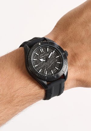 Fekete Nautica karóra texturált számlappal és fehér jelzésekkel, a bal csuklón fekete szilikon szíjjal viselve.