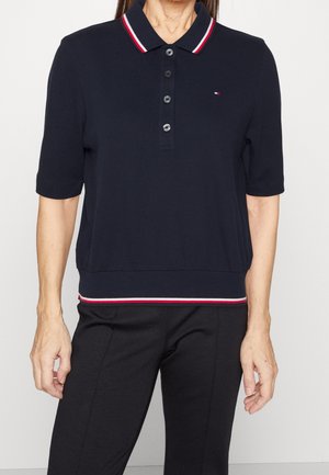 Kobieta ubrana w granatową koszulkę polo z krótkim rękawem, z czerwono-białym paskowanym kołnierzykiem i wykończeniem, w połączeniu z czarnymi spodniami.