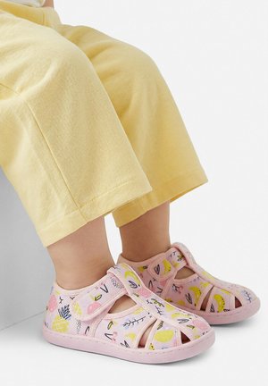 Kind trägt gelbe Hose und pinke Sandalen mit Obst- und Blattmustern und steht vor einem weißen Hintergrund.