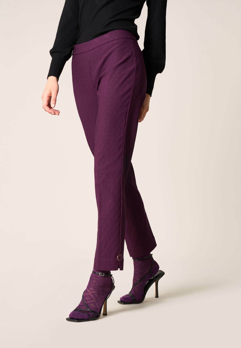 Femme portant un pantalon slim violet coupé court avec une fente latérale et des talons hauts noirs à brides sur des chaussettes violettes à motifs, sur un fond uni.