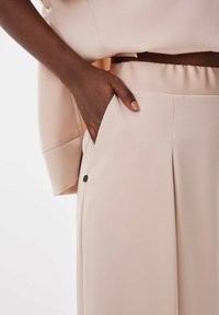 Beige, vloeiende broek met zijzakken en zichtbare stiksels. Een zwarte knoopaccent op de zak en een gladde stoftextuur.