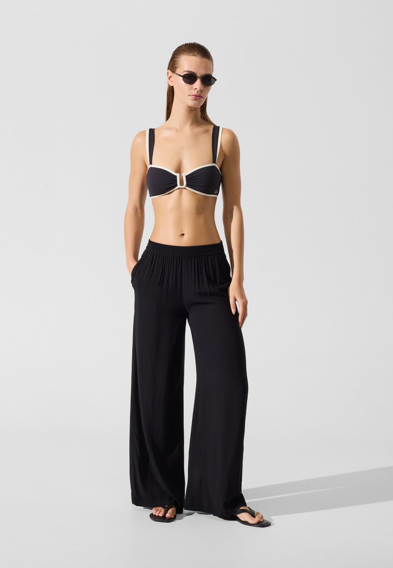 Maillot de bain deux pièces noir avec un accent crème, haut style bandeau, et pantalon noir large taille haute ; modèle portant des lunettes de soleil rondes noires.