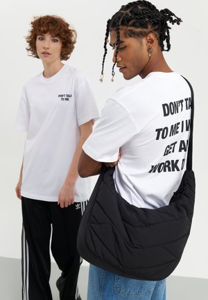 Kobieta z kręconymi włosami i mężczyzna z plecionymi włosami noszą białe koszule z napisem „NIE MÓW DO MNIE”; mężczyzna niesie dużą czarną torbę na ramieniu.