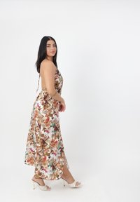Robe maxi florale en beige et marron avec des accents verts ; possède des bretelles spaghetti, une fente sur le côté et une coupe fluide. Portée avec des sandales à talons blanches.