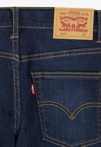 Jeans de mezclilla azul oscuro con una etiqueta de cuero marrón con detalles en rojo y amarillo. Costuras amarillas notables y un diseño distintivo en el bolsillo trasero.