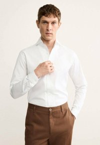 Chemise blanche à manches longues avec un col boutonné, associée à un pantalon marron. La chemise présente une texture lisse et une coupe ajustée.