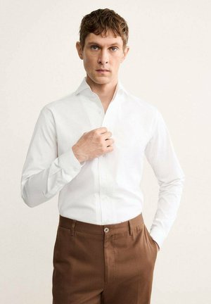 Camicia bianca a maniche lunghe con colletto button-down, abbinata a pantaloni marroni. La camicia presenta una texture liscia e un design aderente.