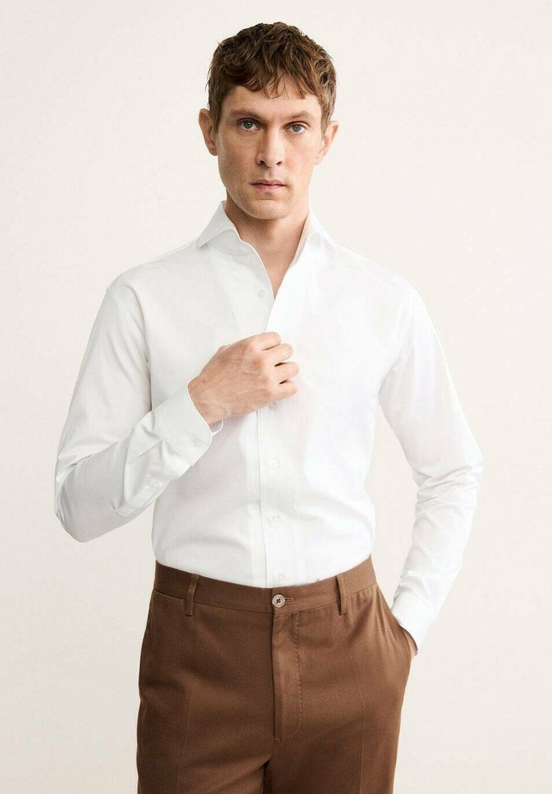 Chemise blanche à manches longues avec un col boutonné, associée à un pantalon marron. La chemise présente une texture lisse et une coupe ajustée.