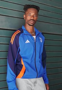 Chaqueta atlética azul y navy con acentos naranjas, que presenta un cierre frontal con cremallera y un logotipo. Hecha de un material suave y ligero.